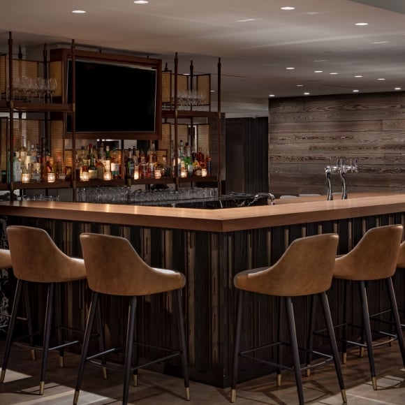 Sleek, modern wooden bar at Revel Lounge featuring tan leather barstools, ambient lighting, and dark marble accents.