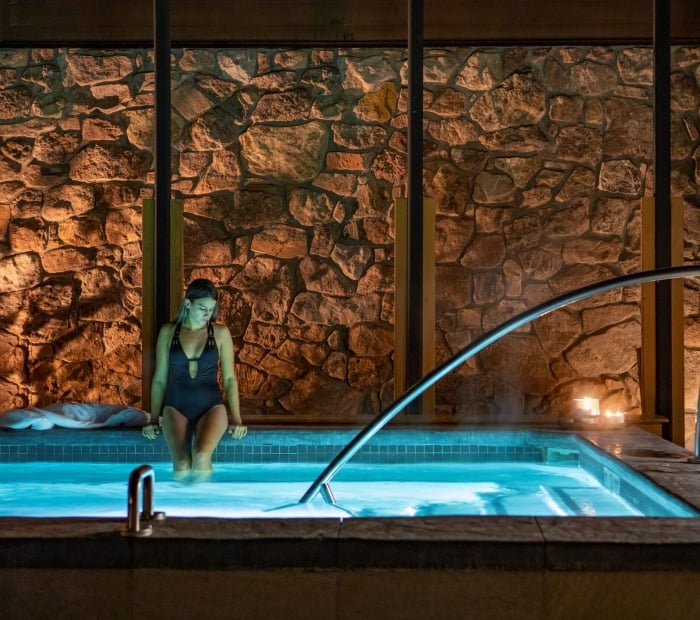 A woman in a black swimsuit sits on the edge of a steaming indoor hot tub set against a rustic stone wall.