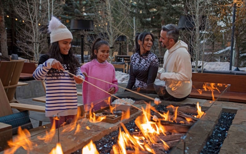 A happy family roasting marshmallows over a linear outdoor fire pit on a cozy, snowy evening at The Hythe.